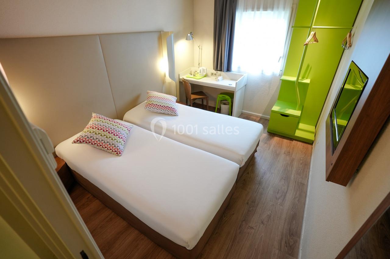 Chambre d'hôtel avec deux lits simples, bureau près d'une fenêtre, armoire verte et sol en parquet.