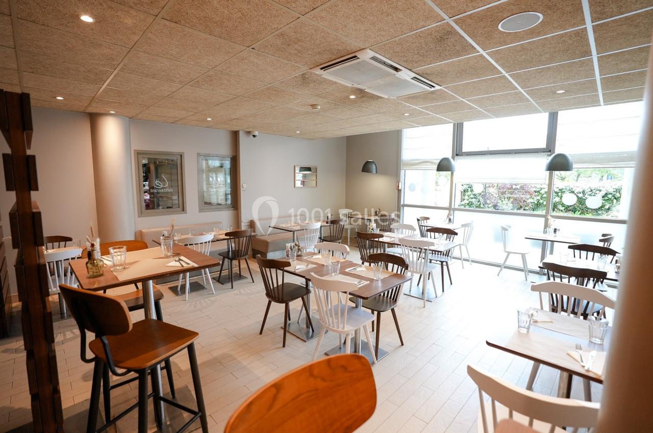 Salle de restaurant lumineuse avec des tables en bois clair, des chaises variées et une grande fenêtre donnant sur l…