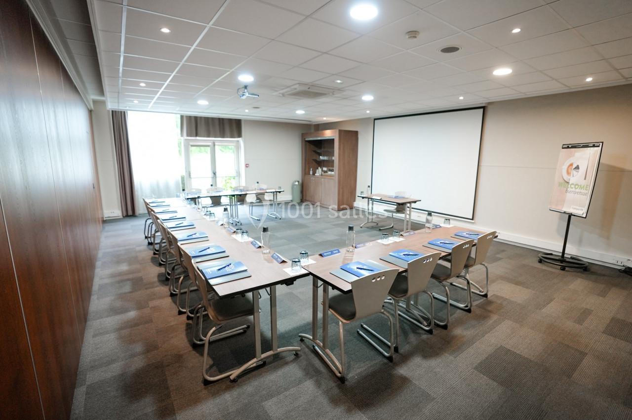 Salle de réunion lumineuse avec tables disposées en U, chaises, écran de projection et tableau de présentation.