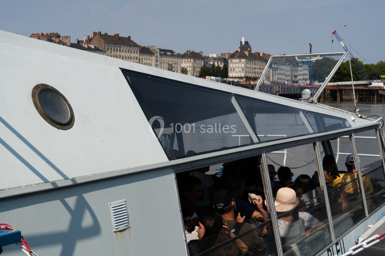 Bateau touristique avec passagers à bord, naviguant sur un fleuve près d'une ville avec des bâtiments en arrière-plan.