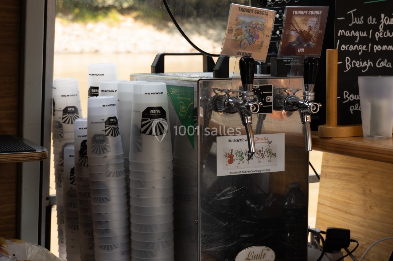 Fontaine à bière avec gobelets réutilisables empilés, posée sur un comptoir en bois près d'un tableau de menu.