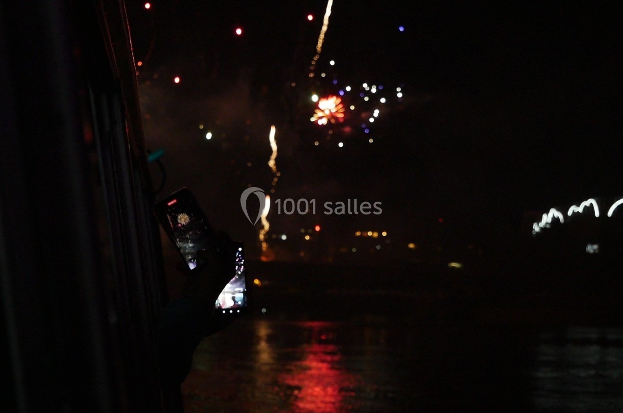 Une personne photographie un feu d'artifice nocturne avec un smartphone, près d'une surface d'eau réfléchissante.