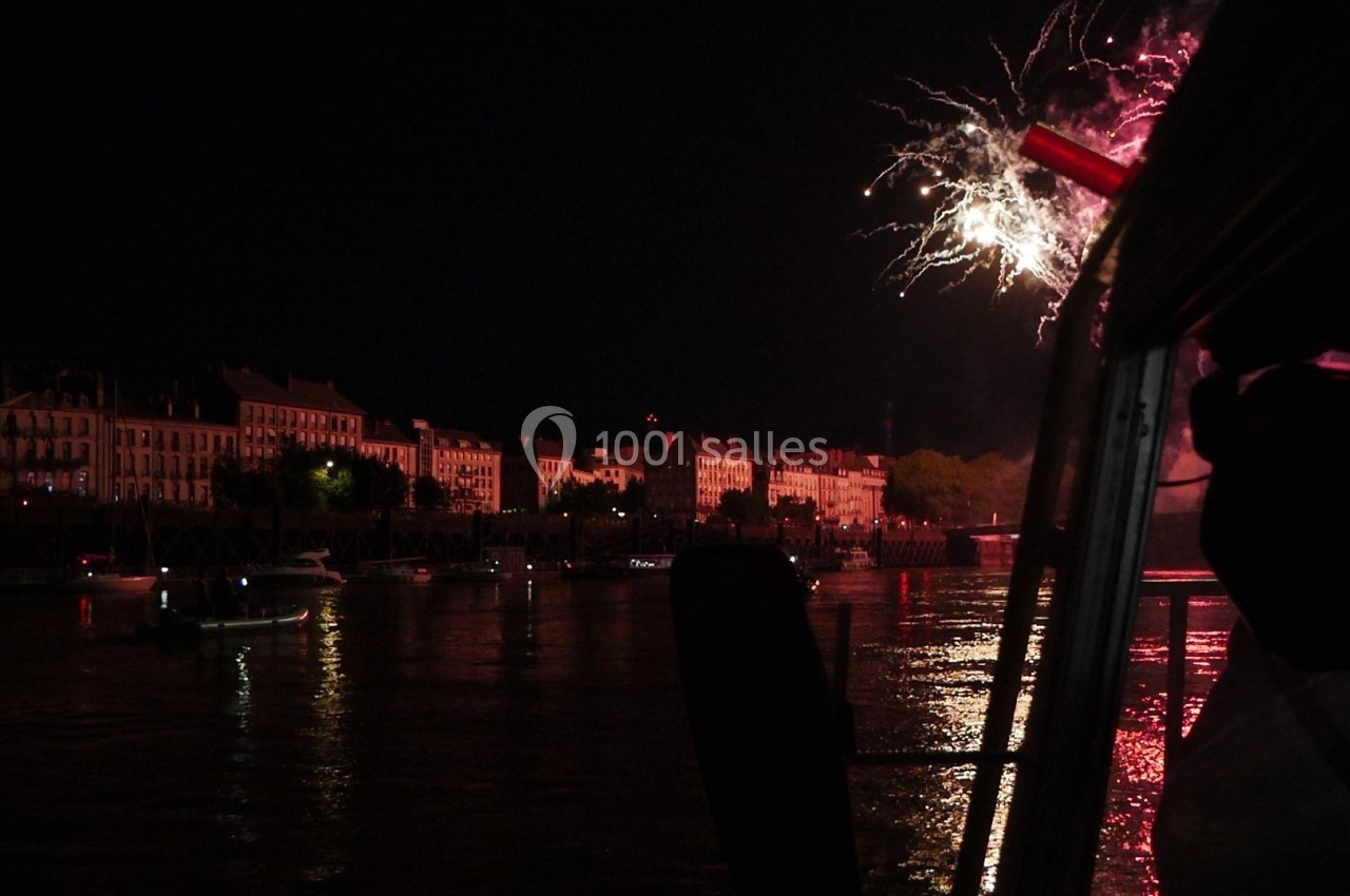 Feu d'artifice illuminant le ciel nocturne au-dessus d'une rivière bordée de bâtiments et de bateaux.