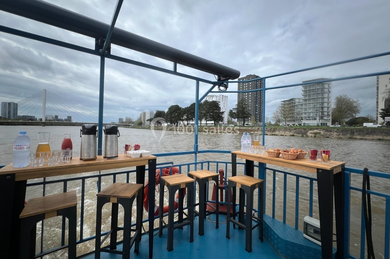 Terrasse d'un bateau avec tables et tabourets, offrant une vue sur un fleuve, des immeubles et un pont en arrière-plan.