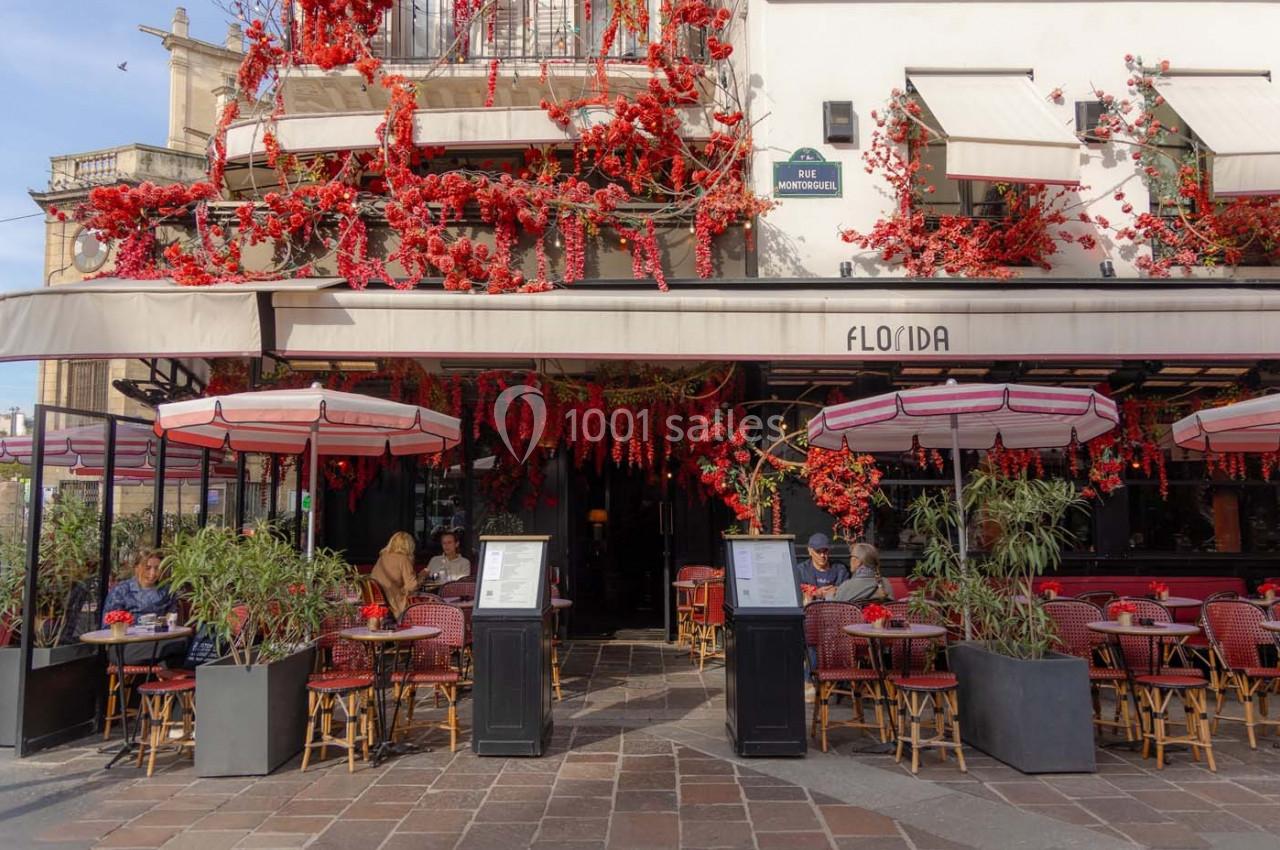 Façade d'un café-restaurant décoré de fleurs rouges, avec terrasse extérieure et tables occupées.
