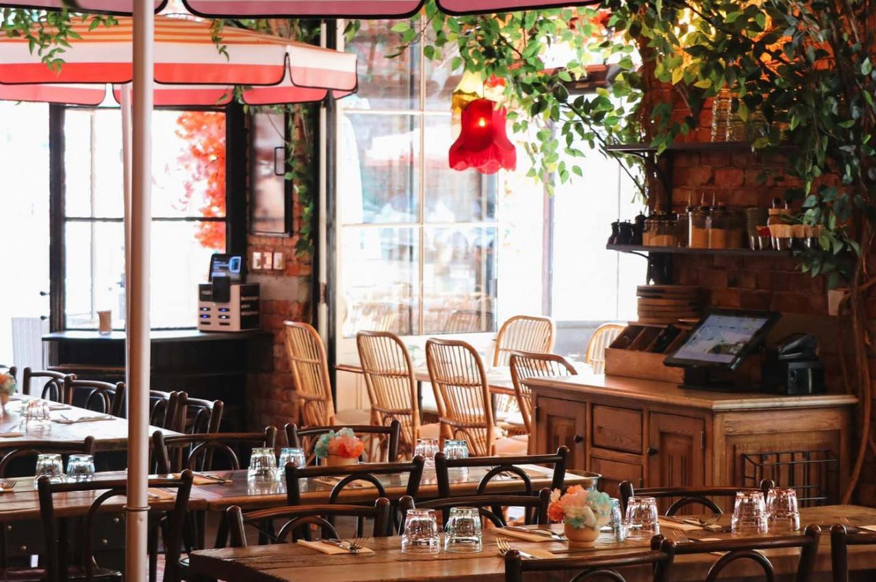 Salle de restaurant lumineuse avec tables en bois, chaises en rotin, plantes suspendues et décoration chaleureuse.
