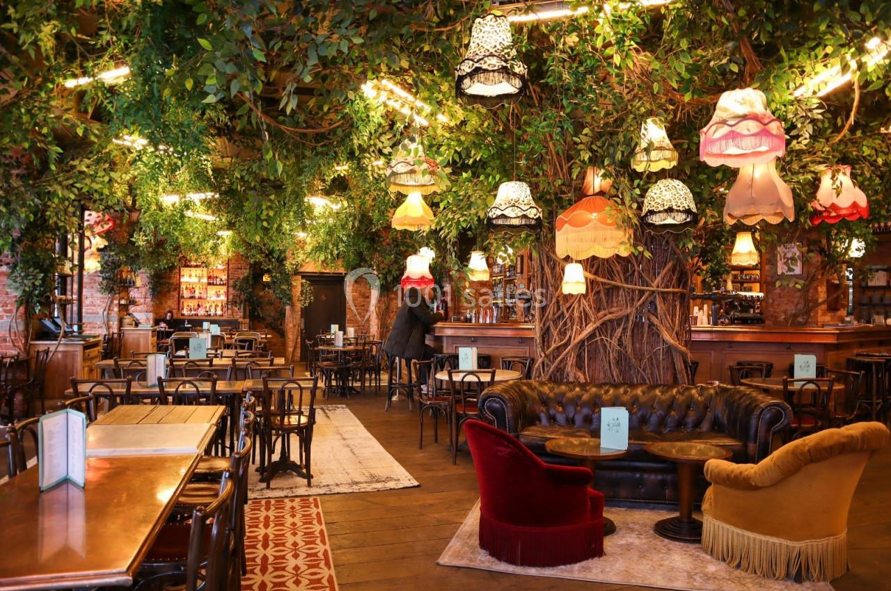 Salle de restaurant décorée avec des plantes suspendues, des lampes vintage et un mobilier éclectique en bois et velours.
