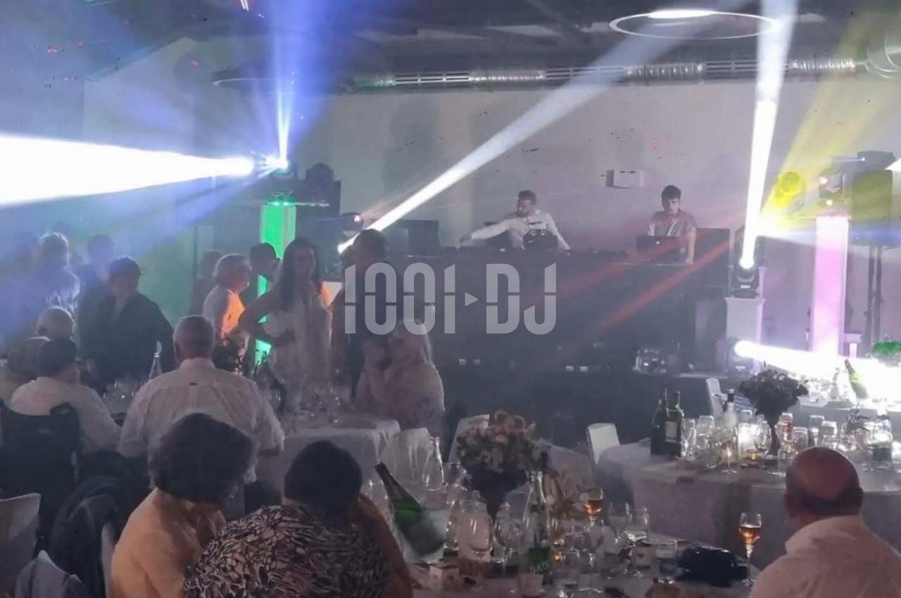 Des convives assis et debout dans une salle de réception éclairée par des jeux de lumière, avec un DJ en arrière-plan.