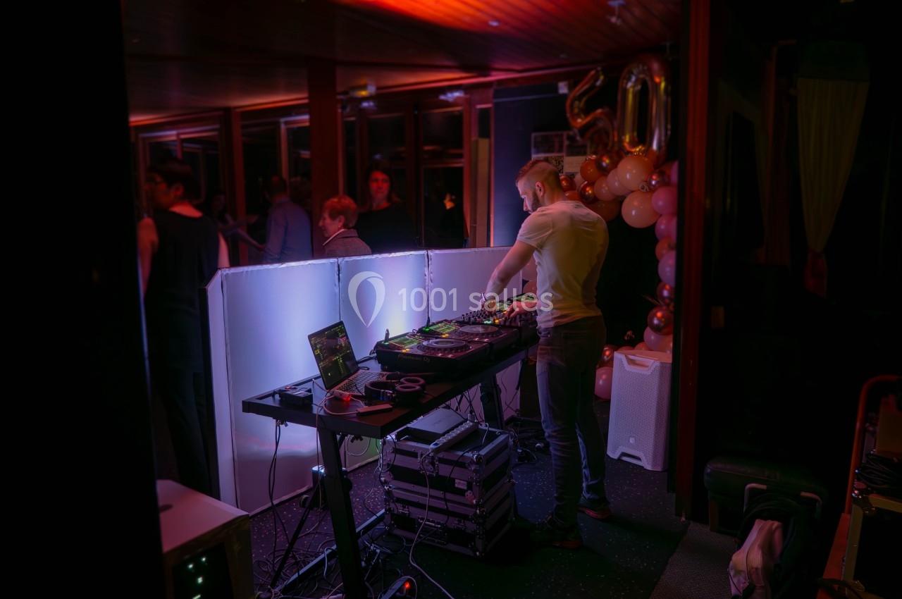 Un DJ mixe de la musique sur une console éclairée dans une salle sombre avec des invités en arrière-plan.