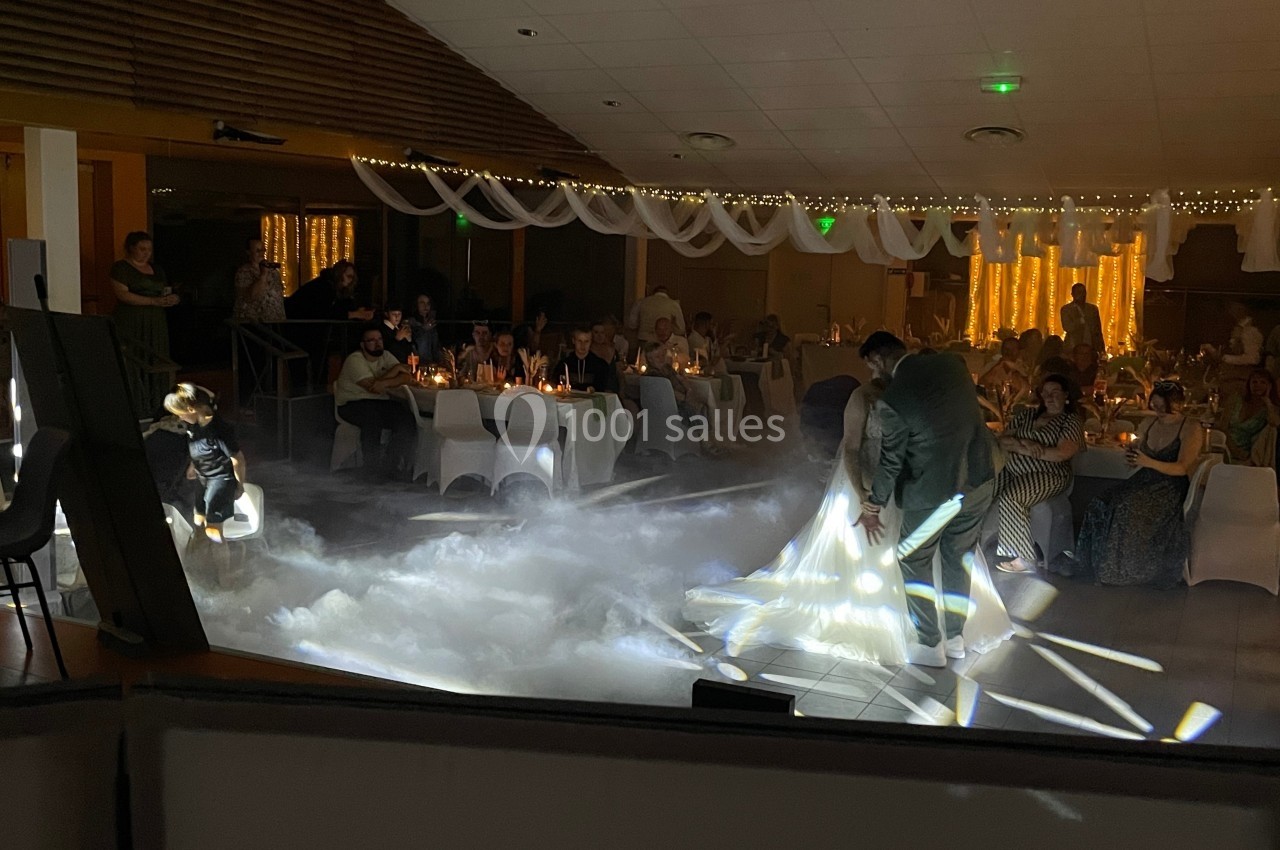 Un couple danse au milieu d'une salle décorée, entouré d'invités assis et d'effets lumineux sur une fumée au sol.