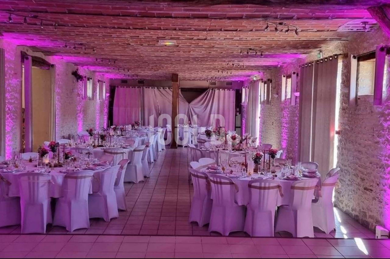Salle de réception décorée pour un mariage, avec tables rondes dressées et éclairage rose sur les murs en pierre.