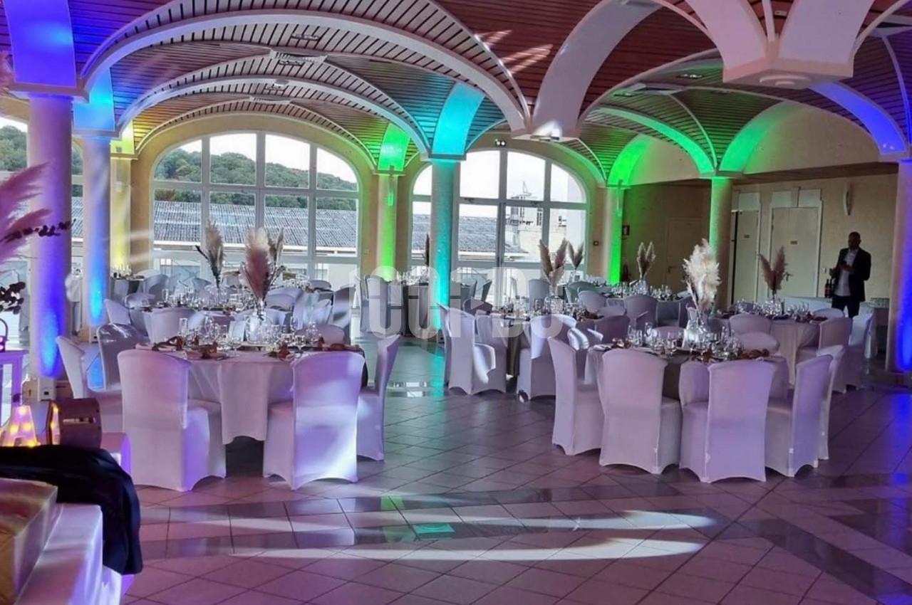 Salle de réception décorée avec des tables rondes, nappes blanches et éclairages colorés, vue sur l'extérieur.
