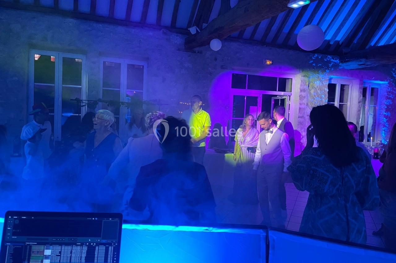 Personnes dansant et discutant dans une salle éclairée par des lumières colorées, avec un DJ en arrière-plan.