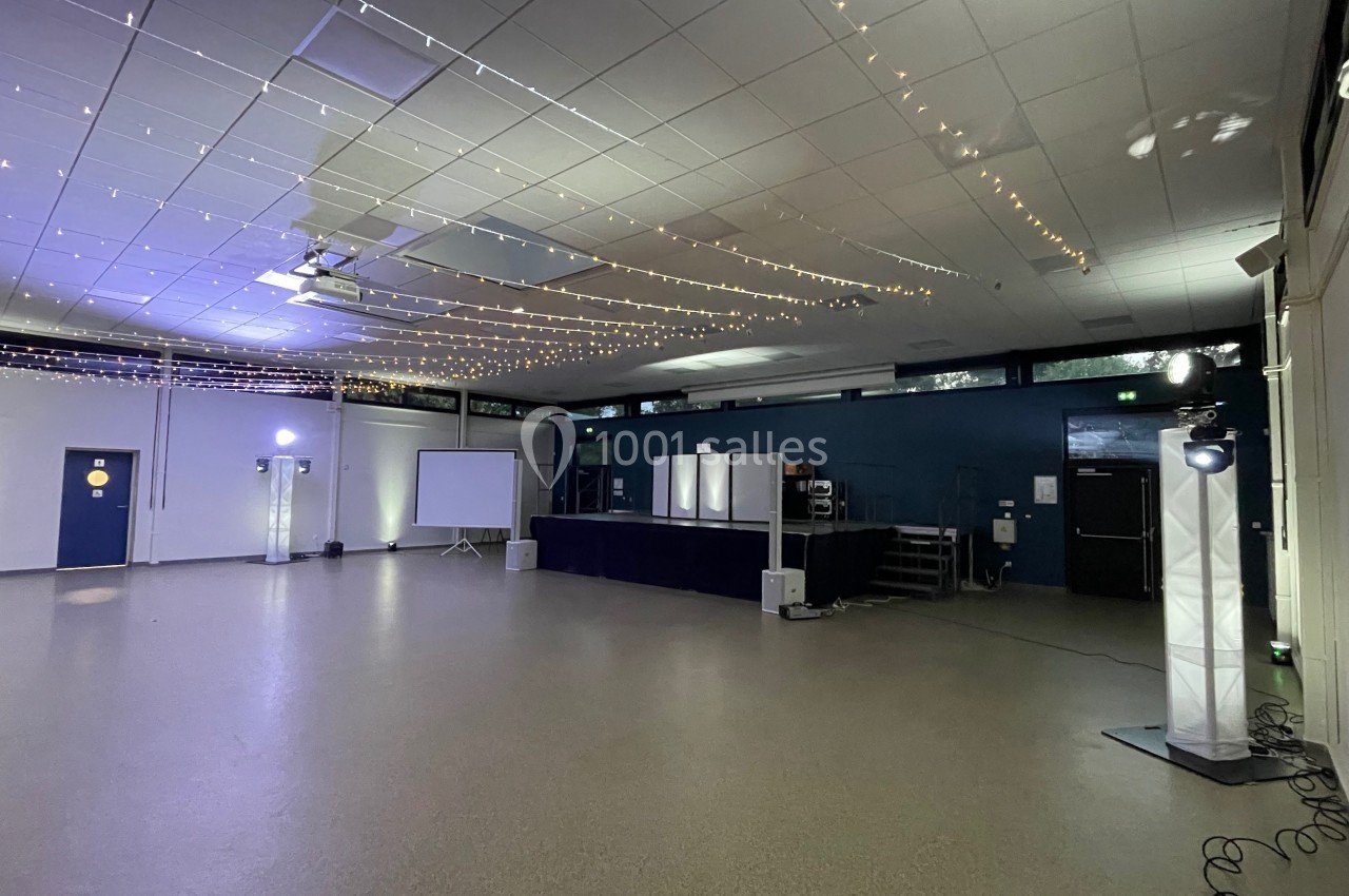 Salle vide avec scène, projecteurs, guirlandes lumineuses au plafond et écran de projection sur le côté.