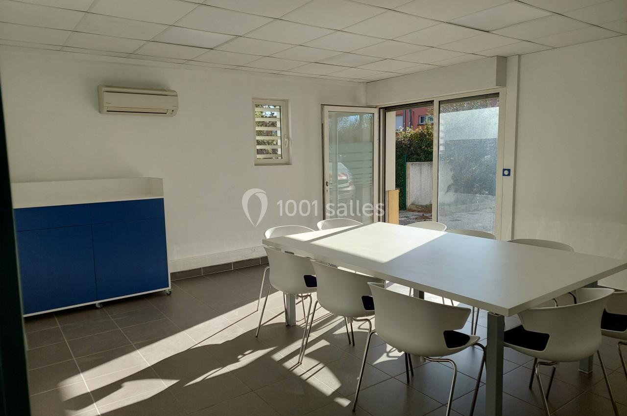 Salle de réunion lumineuse avec table blanche, chaises modernes, meuble bleu et grande baie vitrée donnant sur l'extérieur.