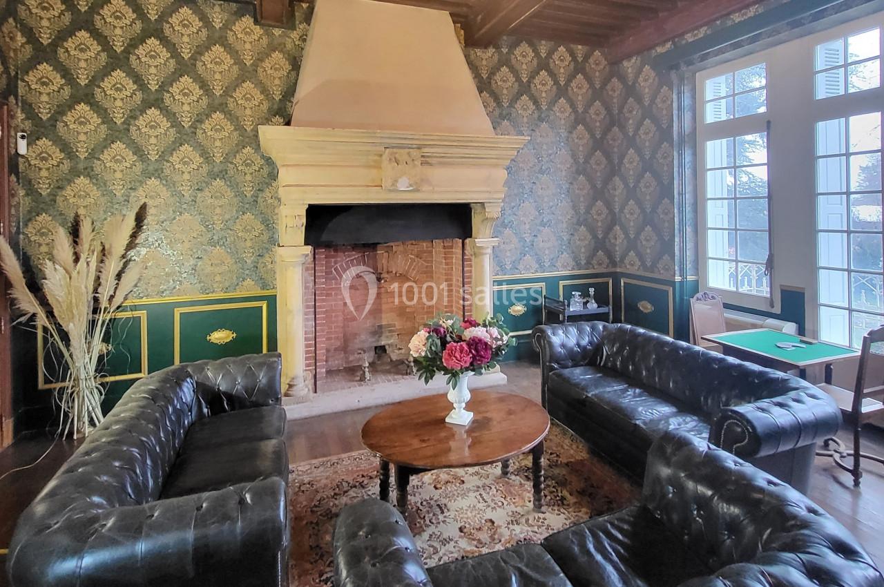 Salon avec cheminée en pierre, murs ornés de papier peint à motifs, mobilier en cuir et table basse avec bouquet de fleurs.