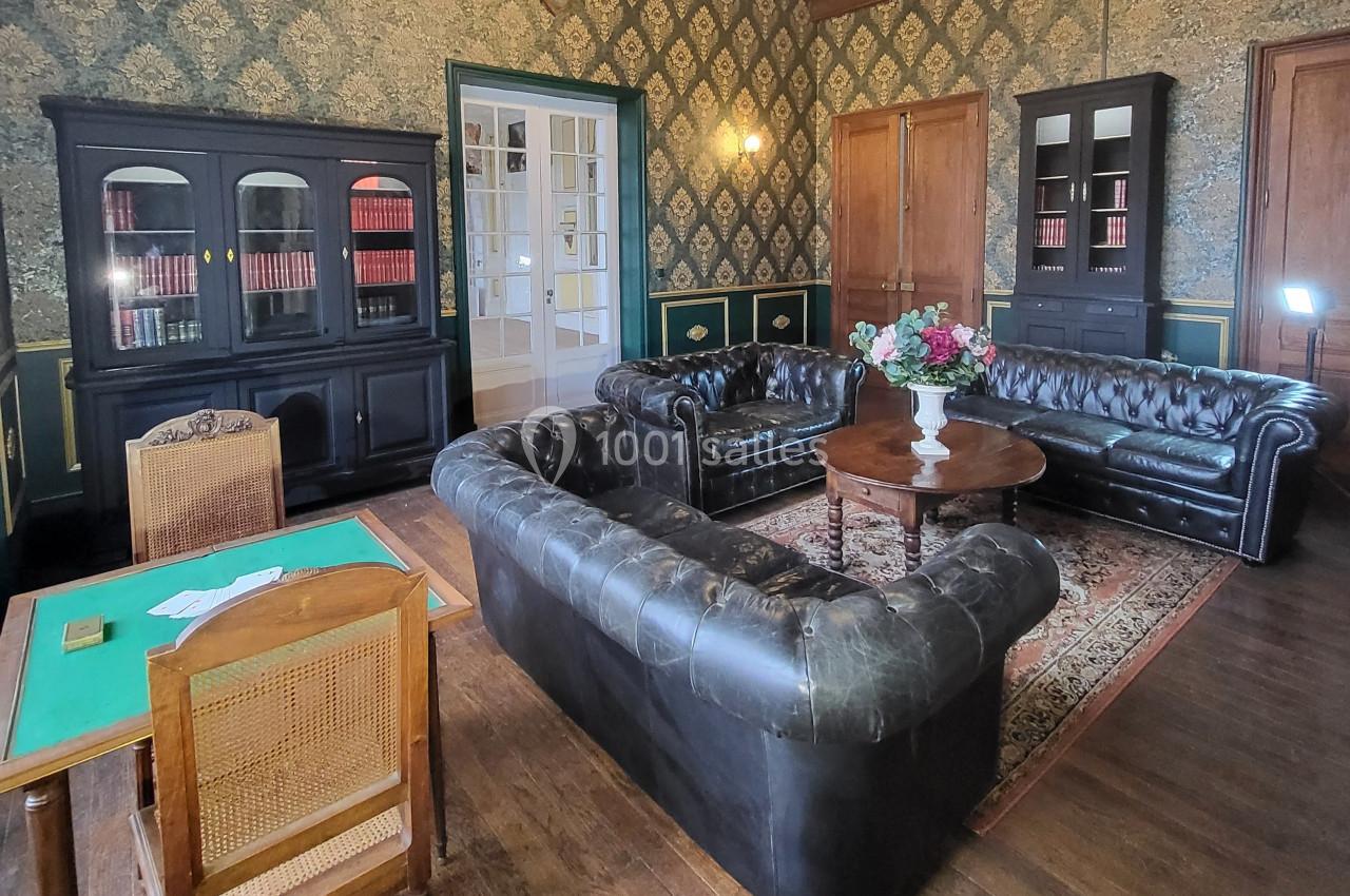Salon avec canapés en cuir noir, table basse ornée de fleurs, bibliothèque et table de jeu sur un parquet en bois.