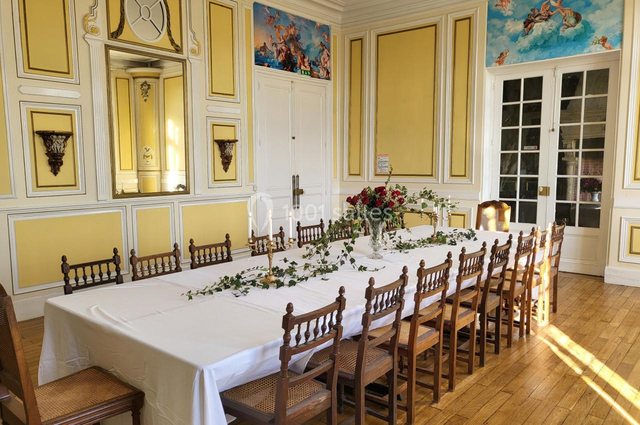 Salle lumineuse avec une grande table rectangulaire décorée de feuillage et de fleurs, entourée de chaises en bois.