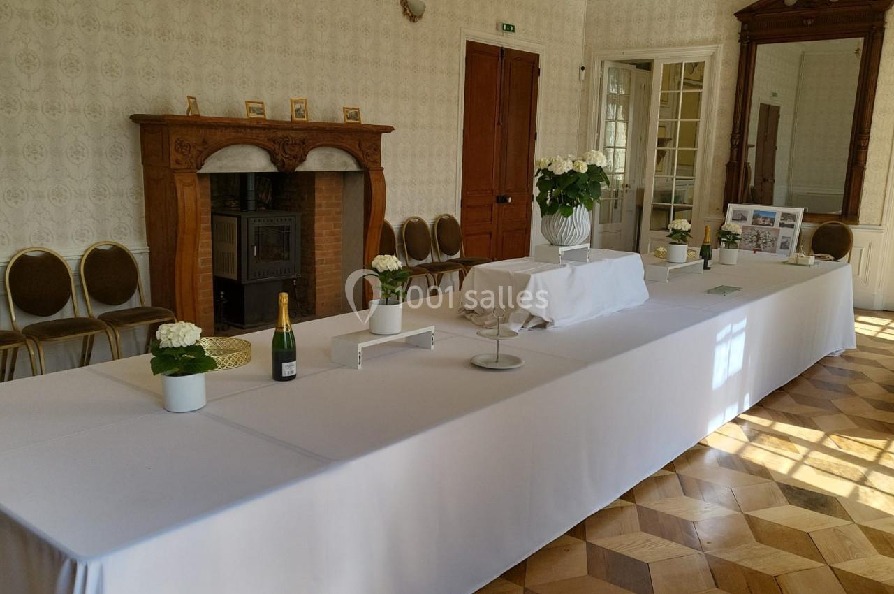 Salle lumineuse avec une grande table recouverte de nappes blanches, décorée de fleurs et d'objets, devant une cheminée.
