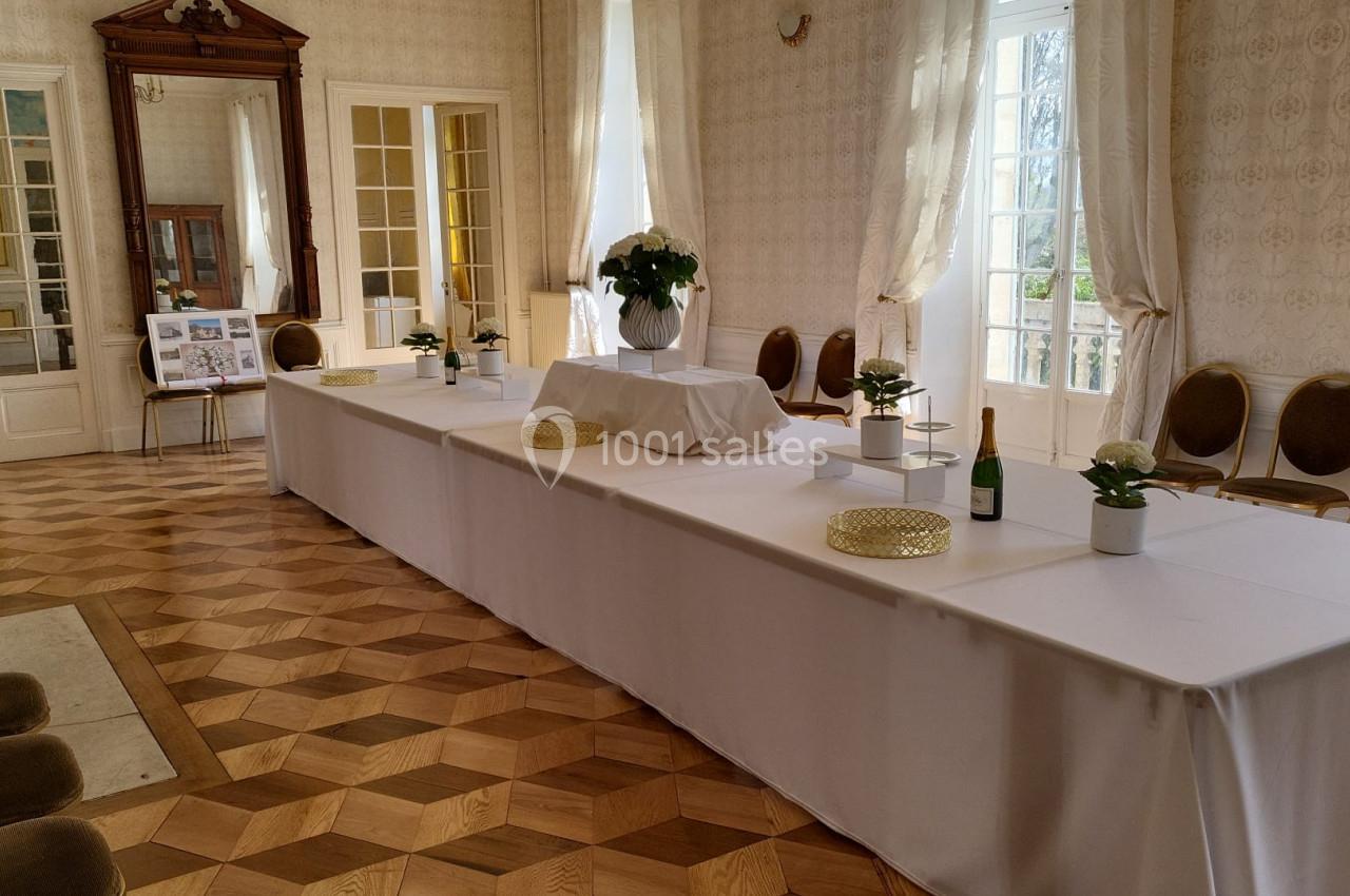 Salle lumineuse avec parquet en bois, tables blanches décorées de plantes et bouteilles, entourées de chaises.