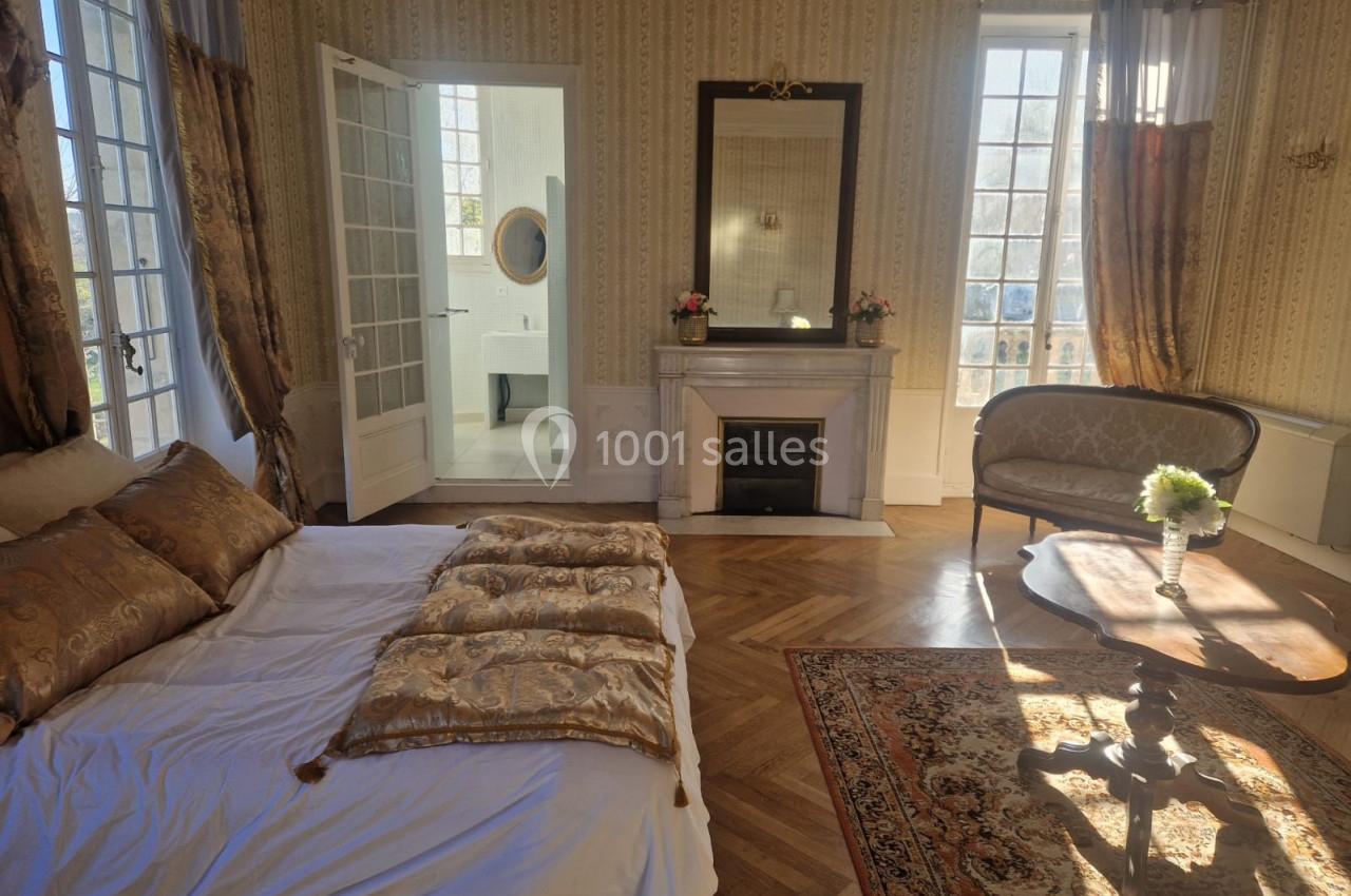 Chambre lumineuse avec lit, cheminée, canapé, table basse et accès à une salle de bain, parquet et tapis au sol.