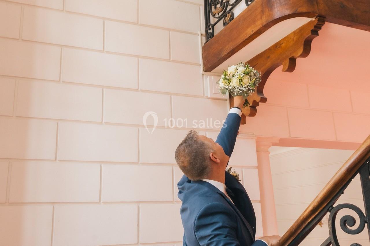 Un homme tend un bouquet de fleurs à une femme souriante sur un balcon, dans un escalier intérieur élégant.