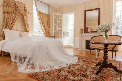 Salon ancien avec parquet en bois, mobilier classique, cheminée, miroir, coiffeuse et grande fenêtre avec rideaux roses.