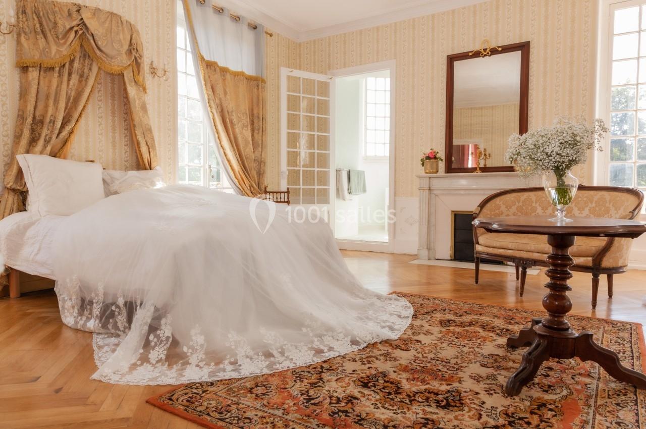 Chambre lumineuse avec lit à baldaquin, tapis coloré, cheminée, fauteuils et rideaux dorés dans un style classique.