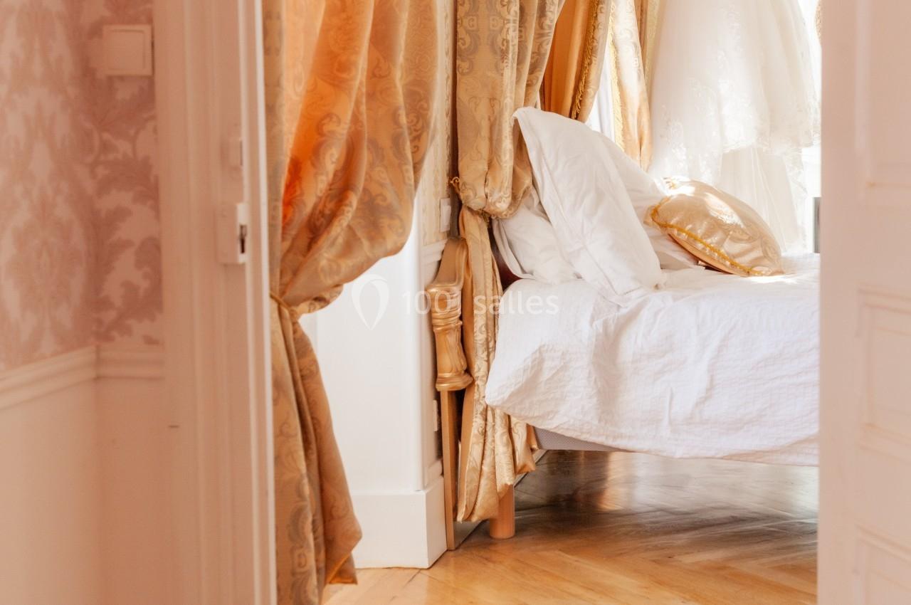 Lit à baldaquin avec rideaux dorés dans une chambre lumineuse, parquet en bois et robe de mariée suspendue en arrière-plan.