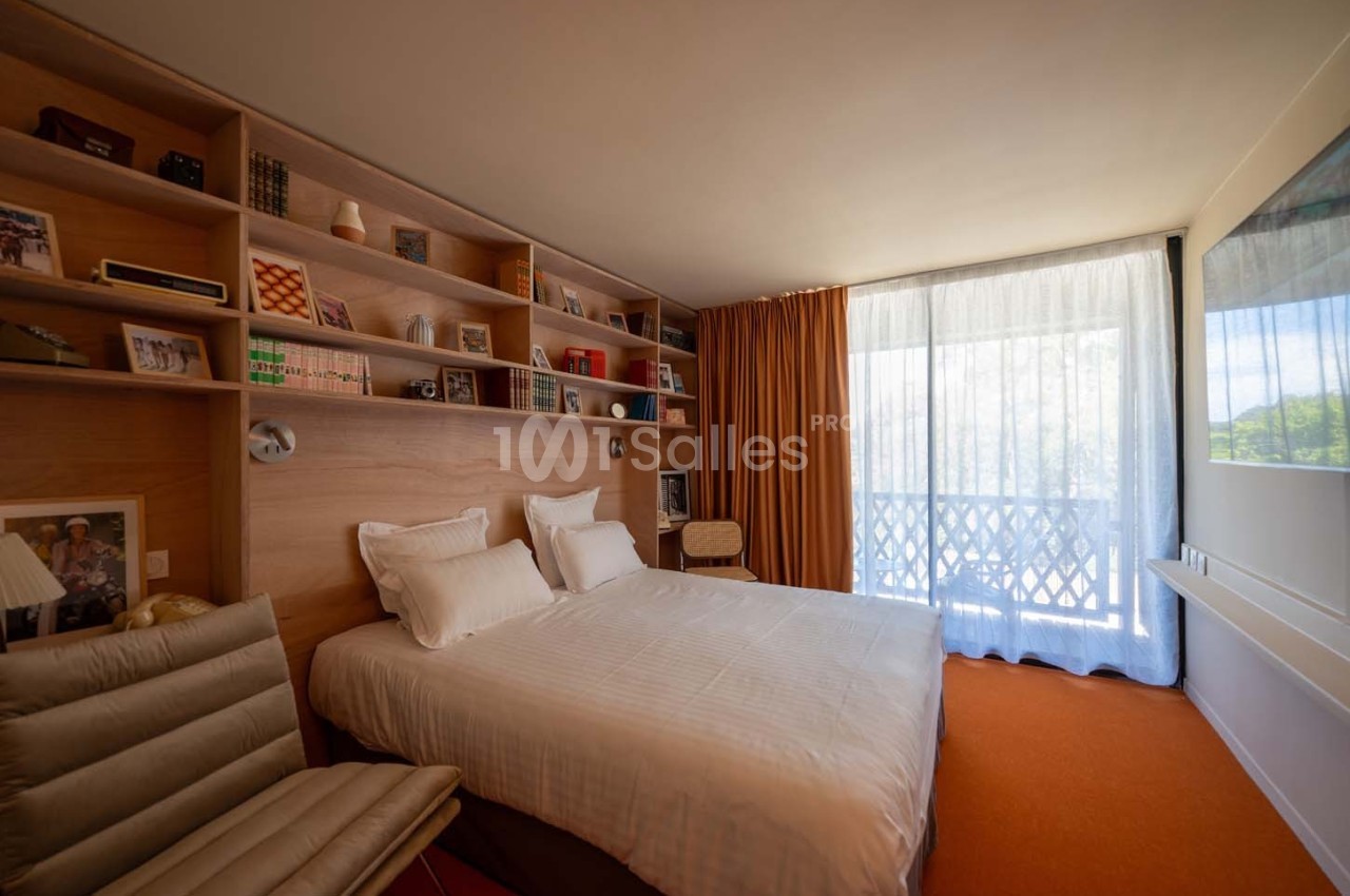 Chambre lumineuse avec lit double, étagères en bois remplies d'objets décoratifs et grande baie vitrée avec rideaux.