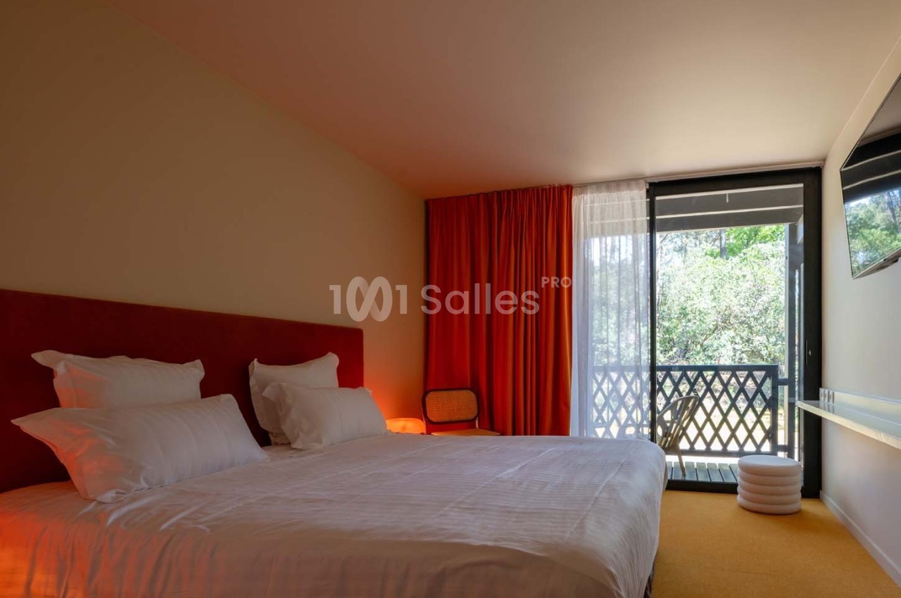 Chambre lumineuse avec lit double, rideaux rouges, balcon donnant sur un espace vert et mobilier minimaliste.