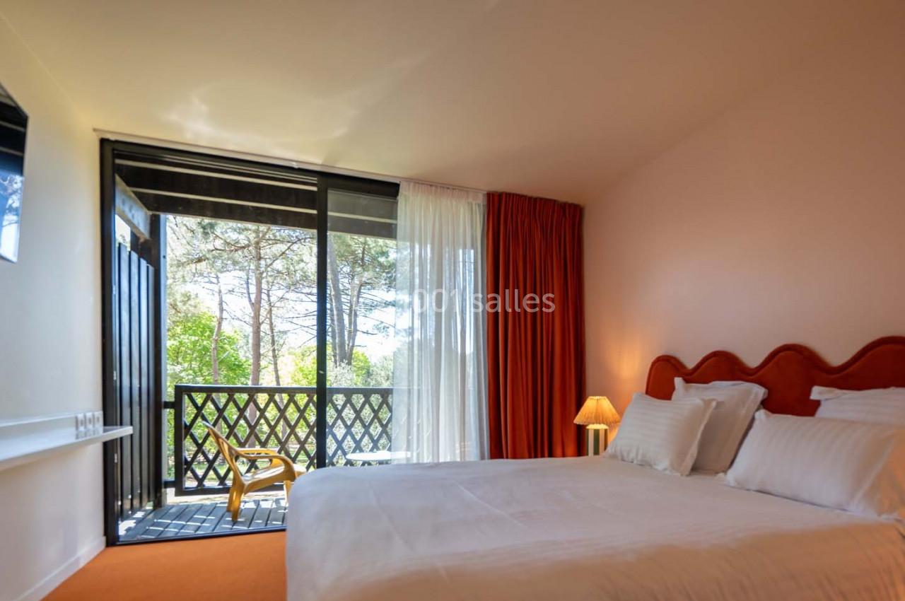 Chambre lumineuse avec lit double, balcon donnant sur un espace arboré, rideaux rouges et lampe de chevet allumée.