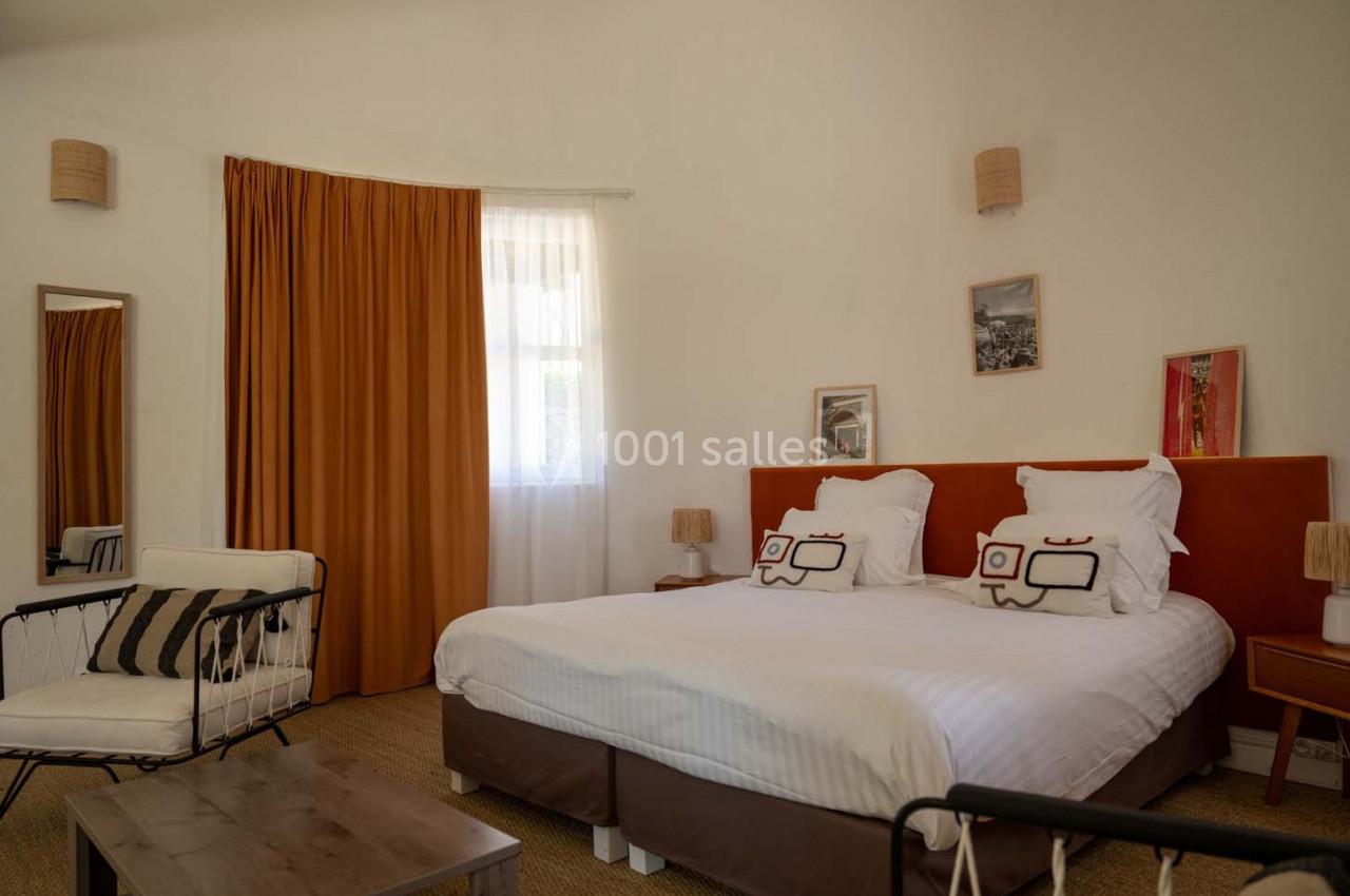 Chambre lumineuse avec un lit double, des coussins décoratifs, un fauteuil, des rideaux orange et des lampes murales.