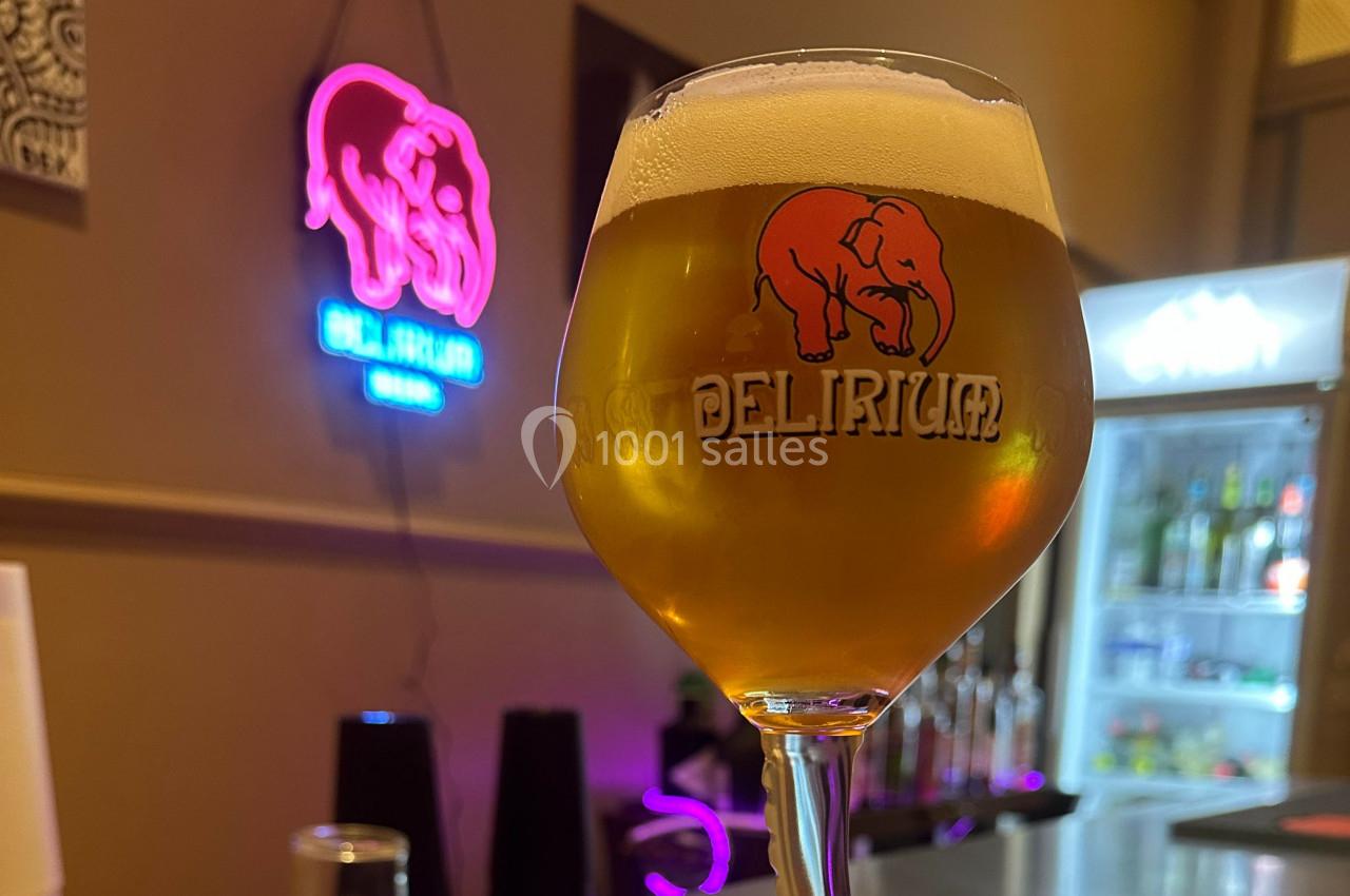Verre de bière blonde Delirium avec mousse, posé devant un néon rose en forme d'éléphant et un réfrigérateur.