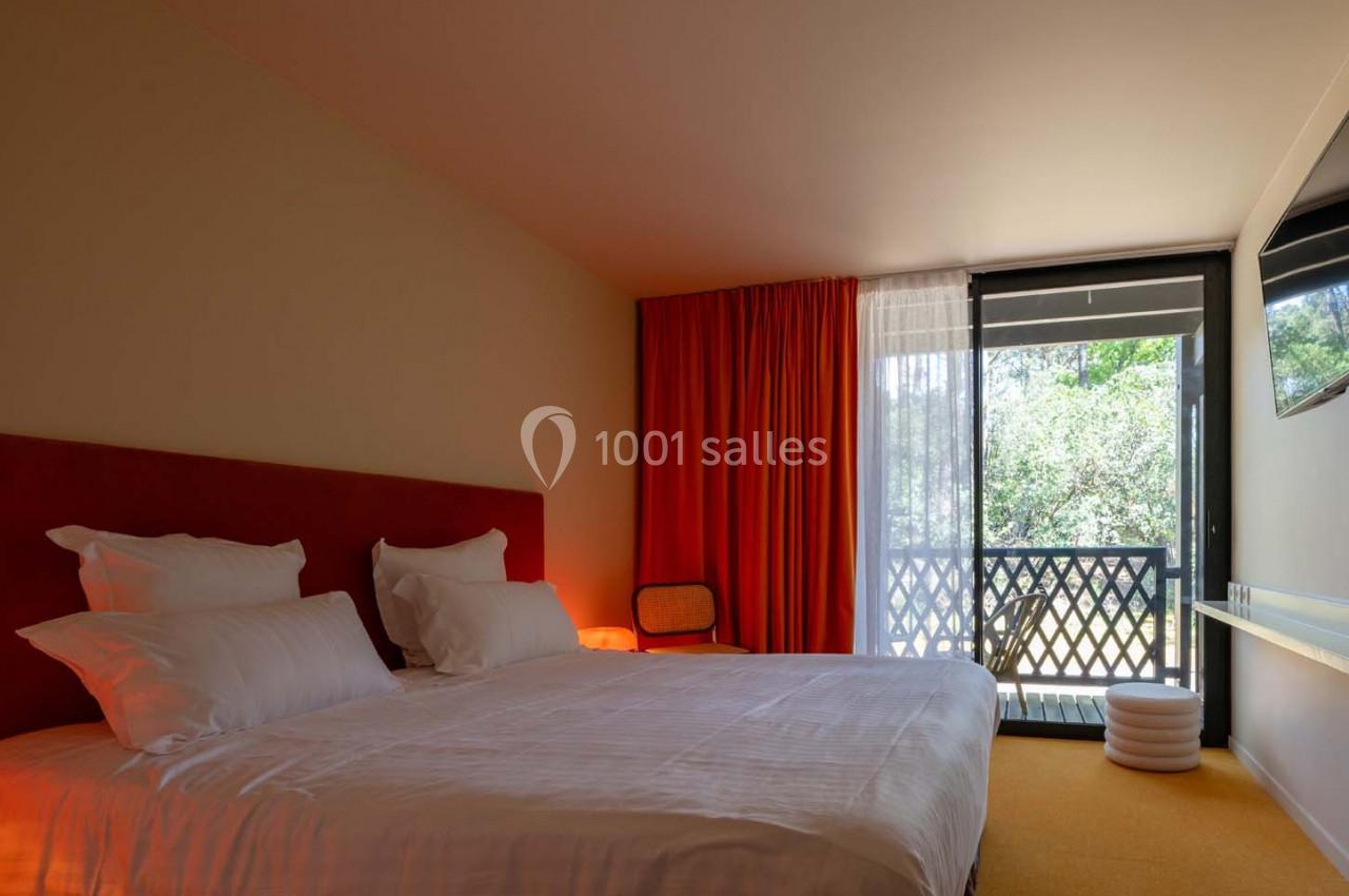 Chambre lumineuse avec lit double, rideaux rouges, balcon donnant sur un espace vert et mobilier minimaliste.