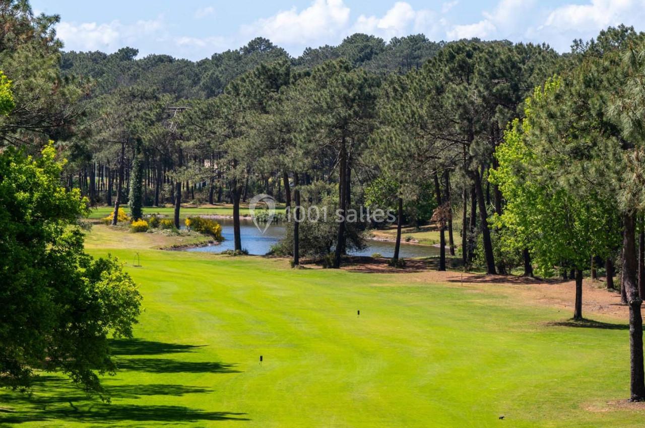 Vue d'un terrain de golf verdoyant entouré de pins, avec un petit étang en arrière-plan sous un ciel dégagé.