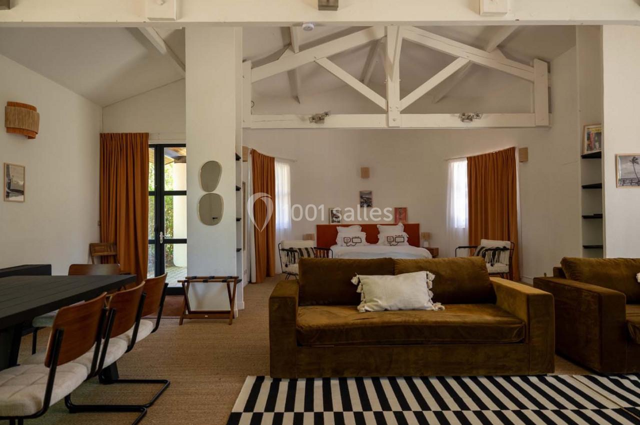 Salon et chambre ouverts avec canapé marron, table à manger, lit double, rideaux orange et poutres apparentes blanches.