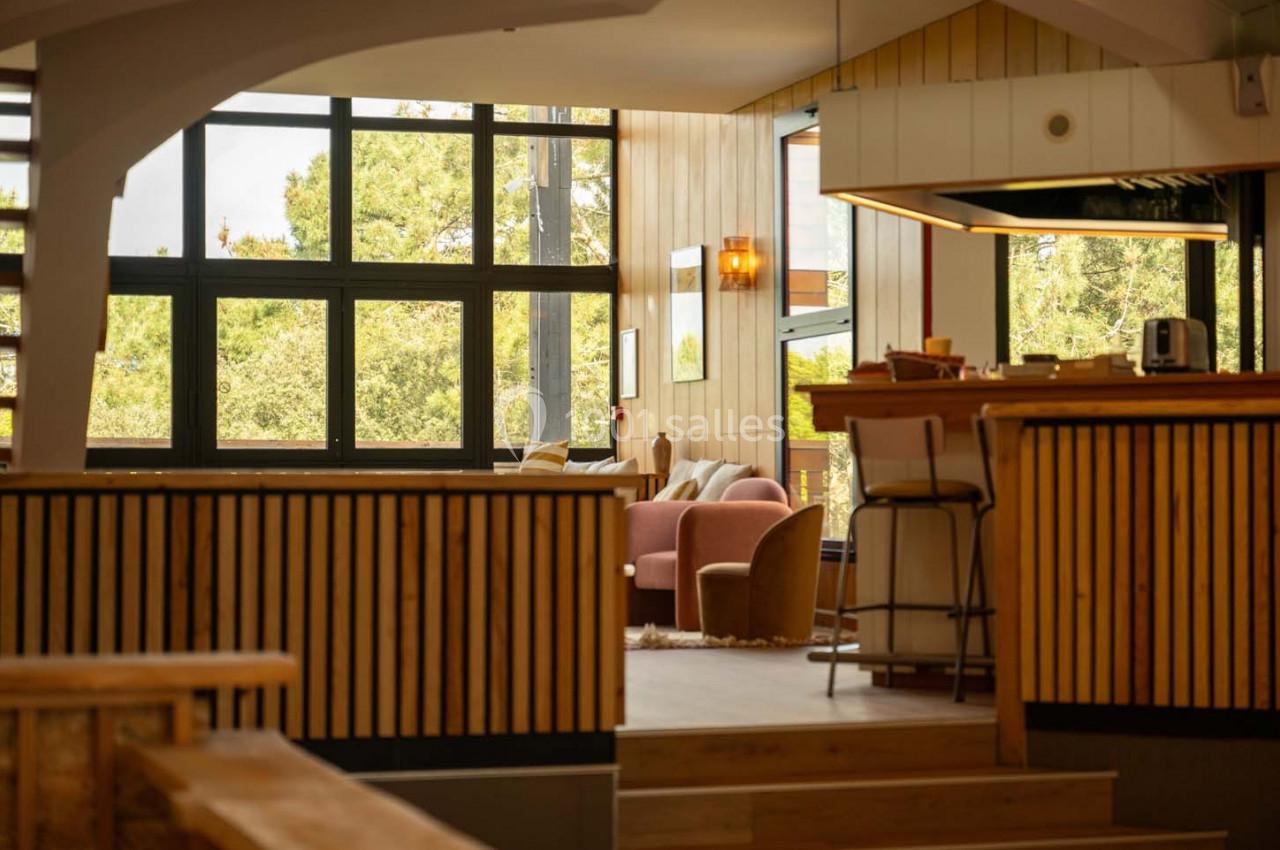 Intérieur lumineux avec grandes fenêtres, mobilier en bois et coin salon cosy avec fauteuils et table basse.