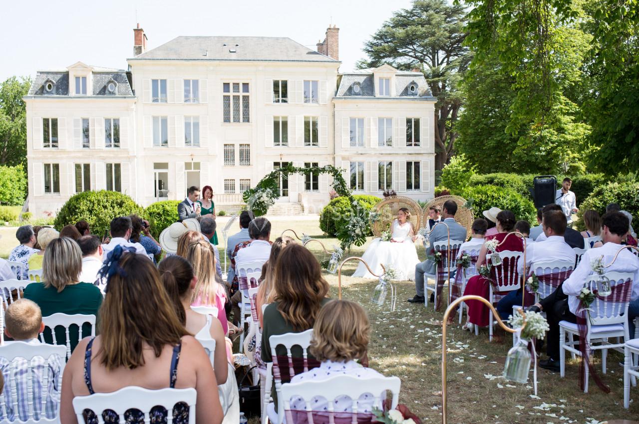 Cérémonie de mariage en extérieur devant un grand bâtiment blanc, avec invités assis et couple au centre.