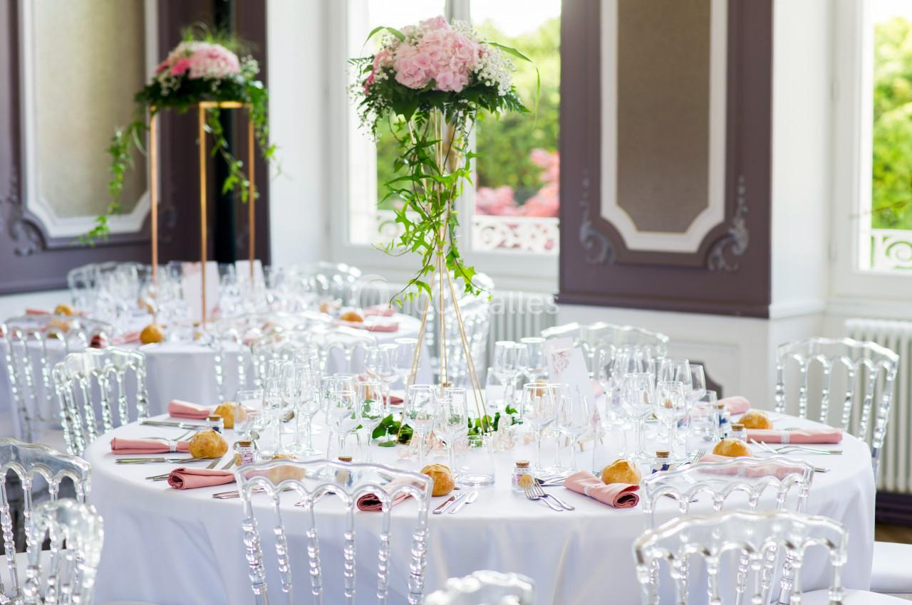 Salle de réception avec tables rondes décorées de nappes blanches, fleurs roses et chaises transparentes.