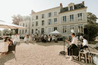 Un groupe de musiciens joue en plein air devant un grand bâtiment de style classique, entouré d'invités en tenue claire.