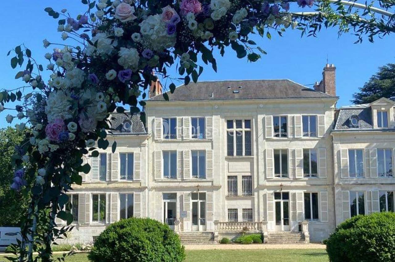 Arche fleurie devant une grande maison de style classique avec façade blanche et jardin verdoyant.