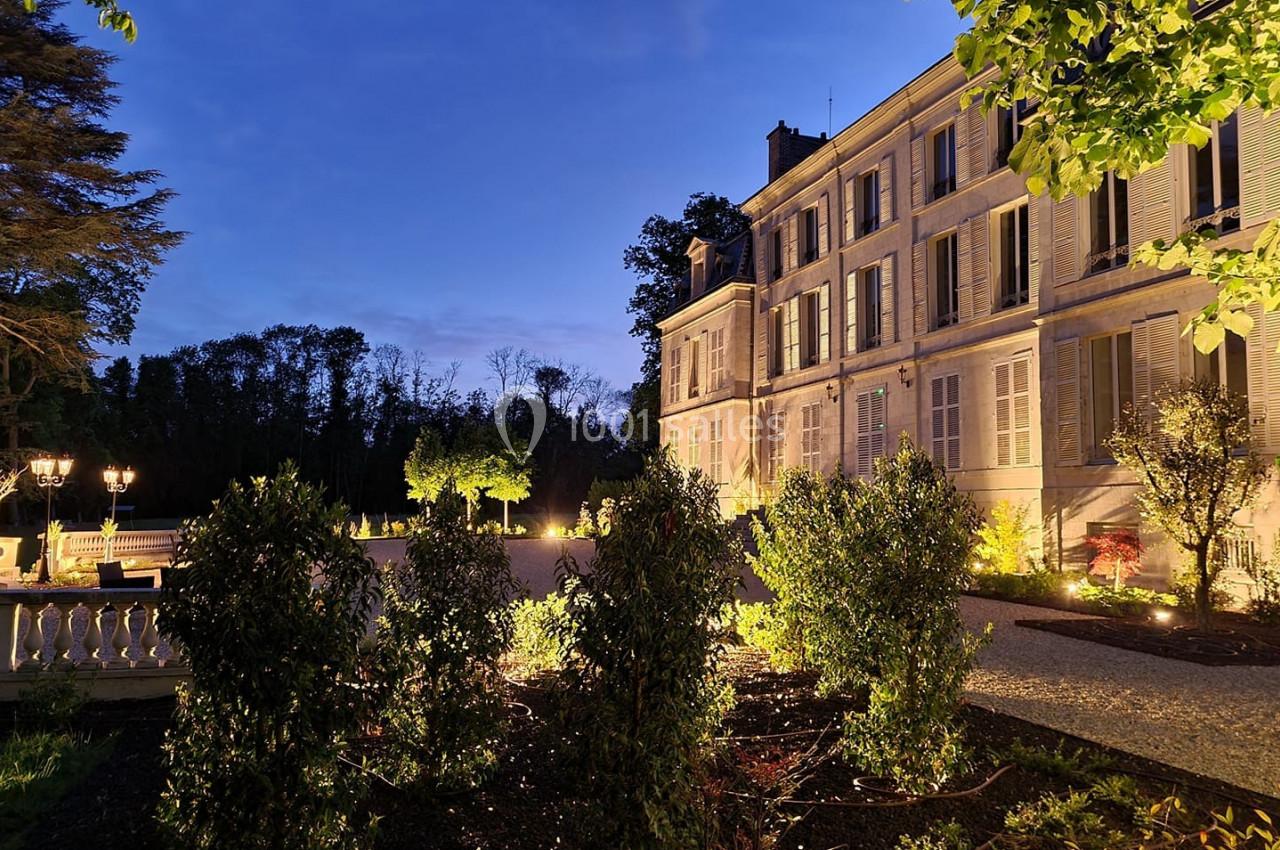 Façade d'un bâtiment élégant éclairé au crépuscule, entouré de végétation et d'une allée gravillonnée.