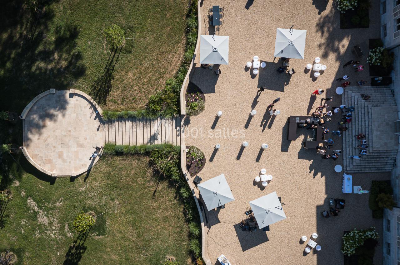 Vue aérienne d'une terrasse avec parasols, tables et invités, entourée de verdure et d'un espace pavé.