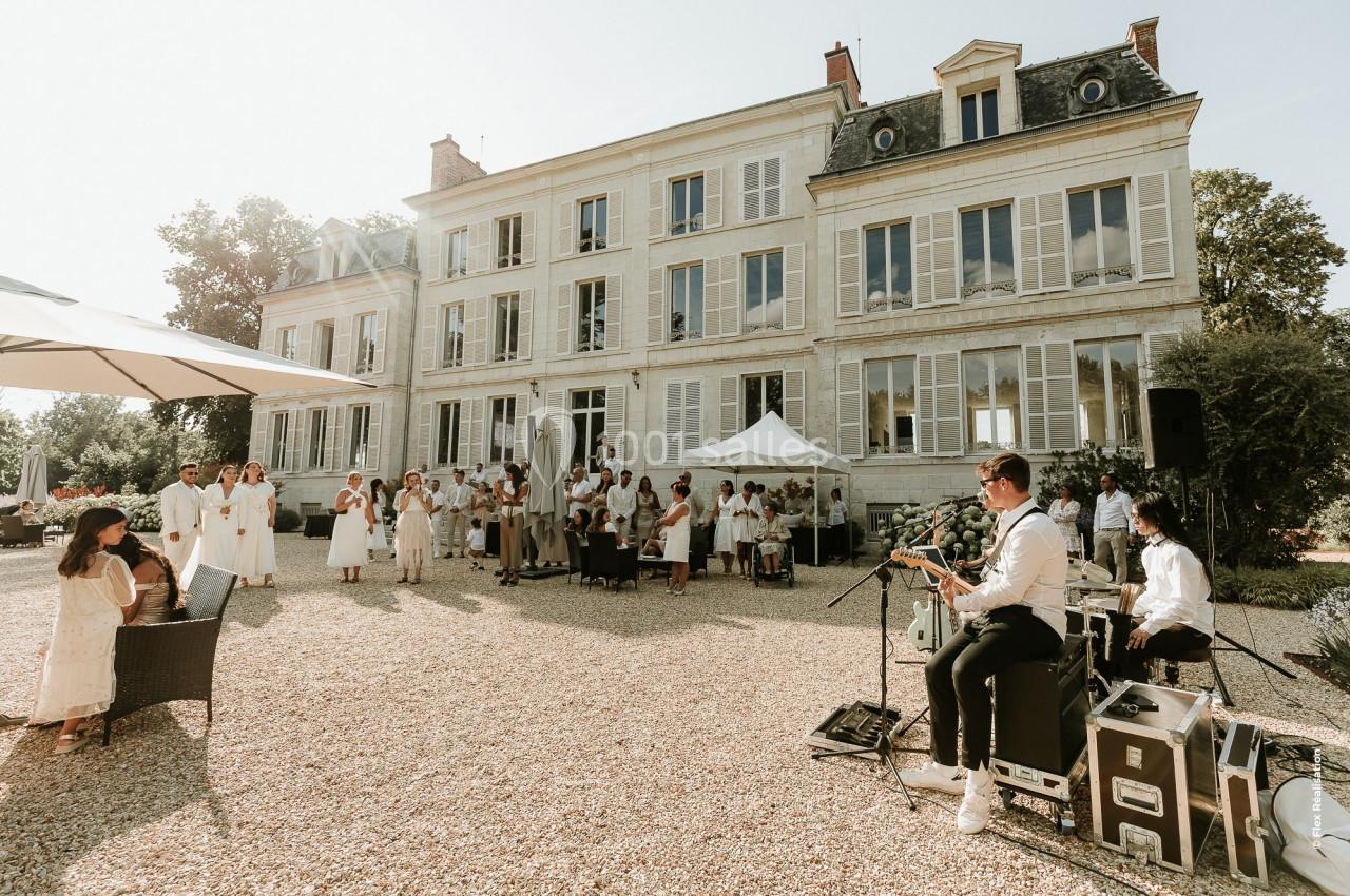 Groupe de musiciens jouant en extérieur devant un manoir, avec des invités en tenue blanche rassemblés autour.