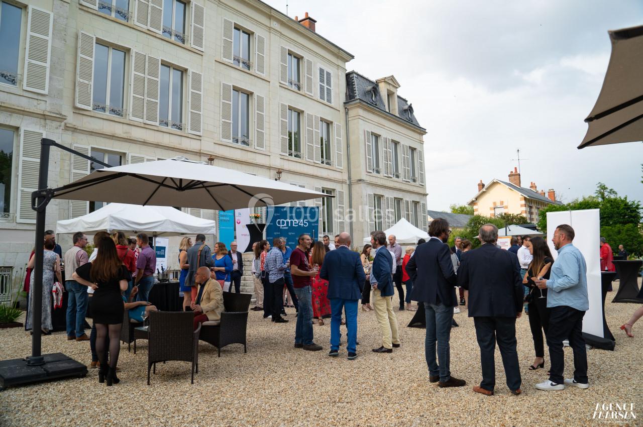 Groupe de personnes discutant dans une cour extérieure devant un bâtiment ancien, sous des parasols et un ciel nuageux.