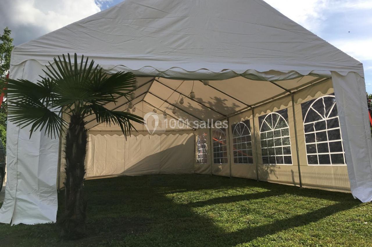 Tente blanche montée sur une pelouse, avec des parois latérales transparentes et un palmier au premier plan.