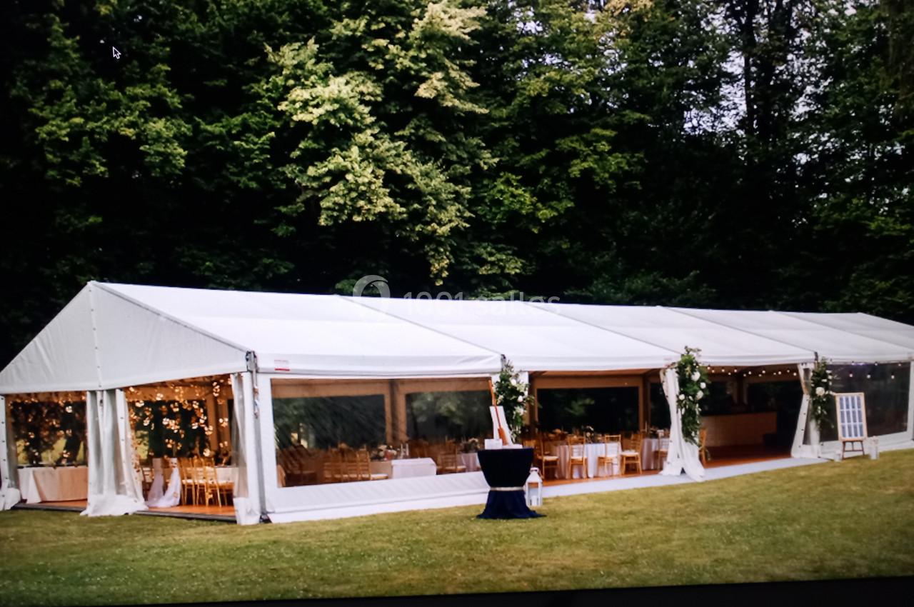 Grande tente blanche installée sur une pelouse, décorée de lumières et entourée d'arbres verdoyants.