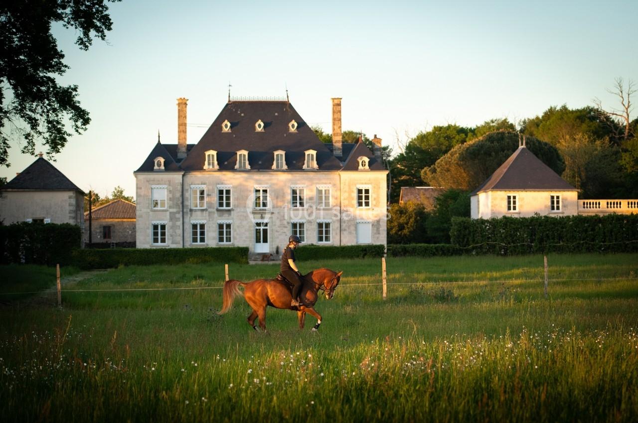 Cavalier montant un cheval dans un champ verdoyant devant un manoir éclairé par une lumière douce en fin de journée.