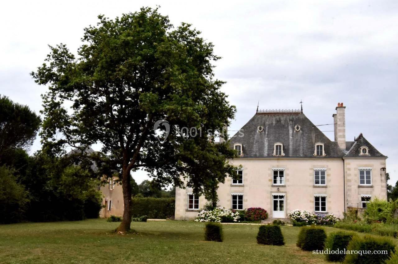 Manoir en pierre blanche avec toit en ardoise, entouré d'un jardin arboré sous un ciel nuageux.